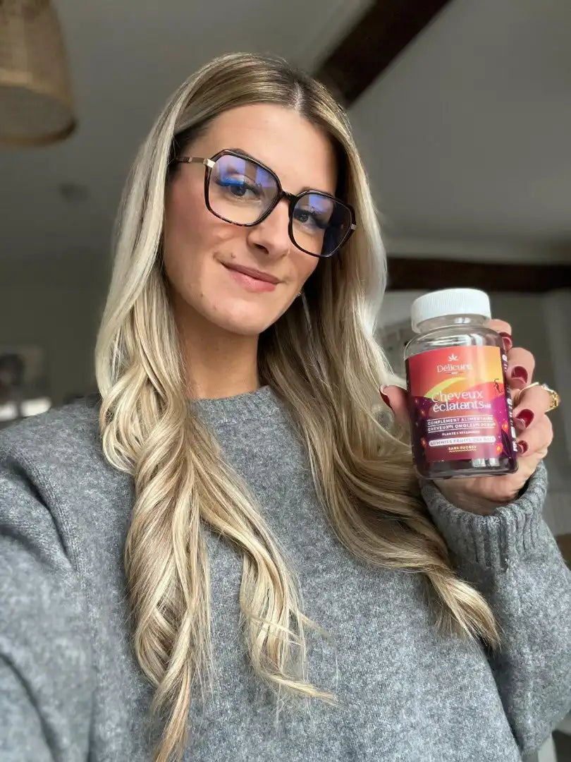 La photo montre une femme blonde souriante portant des lunettes et un pull gris, tenant un flacon de gummies Cheveux Éclatants. Elle présente le produit en soulignant ses bienfaits pour des cheveux plus sains et brillants. La pièce en arrière-plan est bien éclairée, avec un décor chaleureux et des poutres apparentes.