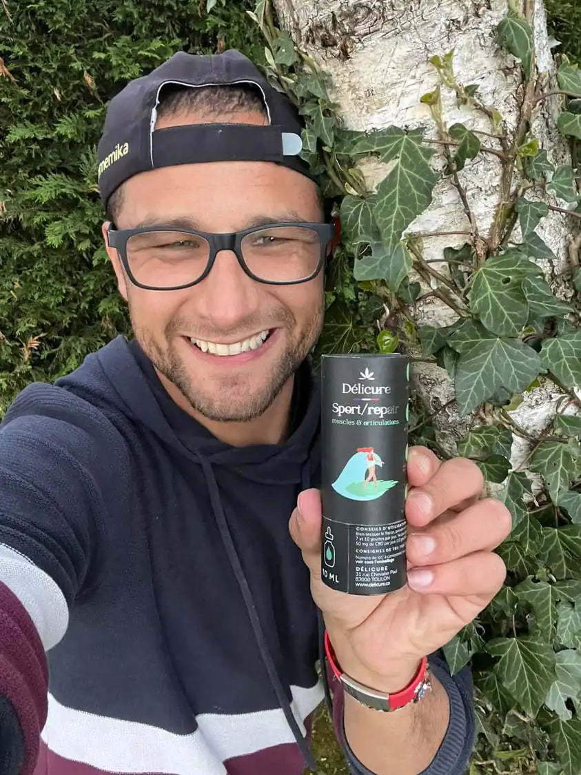 La photo montre un homme souriant, portant une casquette et des lunettes, tenant un flacon de CBD Sport & Repair de la marque Délicure devant un arbre. Il met en avant les bienfaits du produit pour les muscles et les articulations. Il porte un sweat à capuche rayé. L'arrière-plan montre une haie verte.