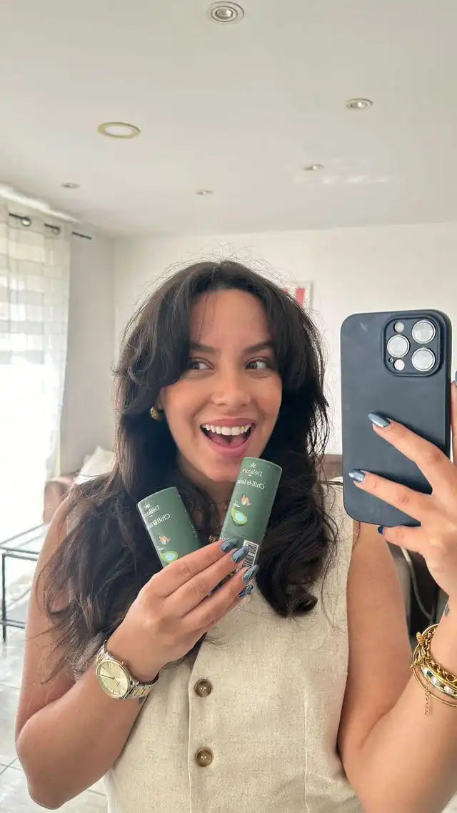 La photo montre une femme souriante tenant deux flacons de CBD Chill & Relax devant un miroir, prenant un selfie. Elle met en avant les bienfaits des produits pour la relaxation et la gestion du stress, dans l'apaisement des douleurs. Elle porte un gilet beige et une montre dorée. La pièce en arrière-plan est lumineuse et moderne.