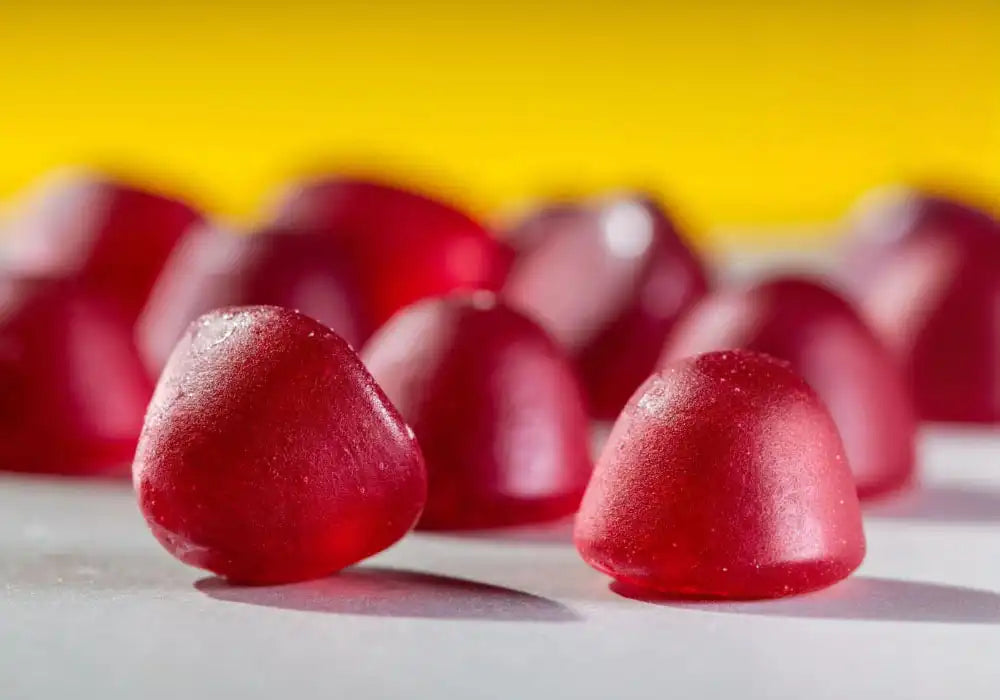 Image de gros plan sur des gummies rouges Délicure Cheveux éclatants, conçus pour améliorer la santé des cheveux. Ces gummies, riches en biotine et autres vitamines, sont spécifiquement formulés pour booster la pousse et la croissance des cheveux. La photo présente plusieurs gummies sur un fond gris avec un arrière-plan jaune vif, mettant en valeur leur texture et leur couleur éclatante.