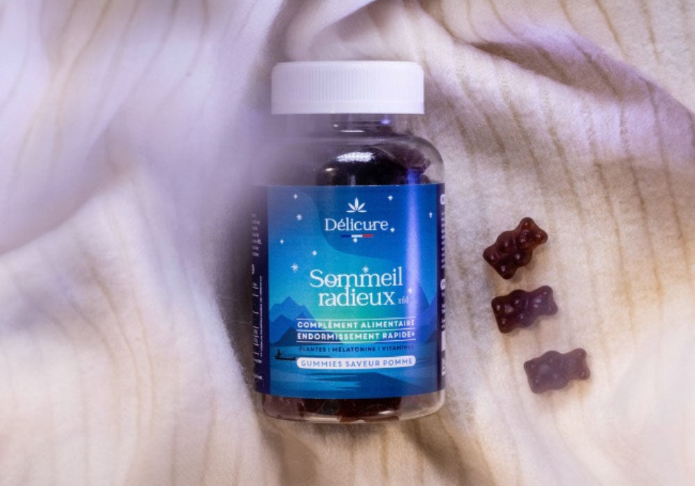 Image d'un flacon de gummies Délicure Sommeil Radieux, conçu pour favoriser un endormissement rapide. Le flacon est posé sur un tissu doux et clair, créant une atmosphère de confort et de relaxation. À côté du flacon, trois gummies en forme d'ourson sont disposés. L'étiquette bleue du flacon présente des illustrations d'étoiles et de montagnes, évoquant une ambiance nocturne et apaisante. Les gummies sont saveur pomme, comme indiqué sur l'étiquette.