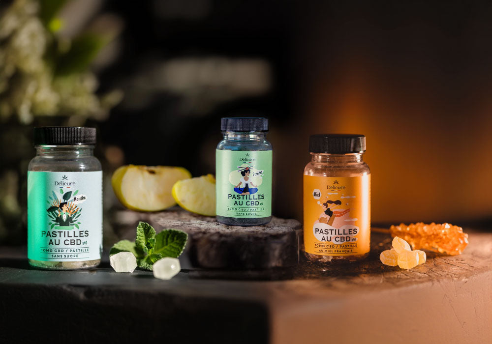 Image de trois flacons de pastilles au CBD de la marque Délicure, disponibles en trois saveurs : menthe, pomme et miel. Chaque flacon contient des pastilles sans sucre, conçues pour aider à gérer le stress et promouvoir la relaxation. La photo montre les flacons disposés sur une surface naturelle, entourés d'ingrédients correspondant à chaque saveur, comme des feuilles de menthe, des tranches de pomme et du miel, mettant en avant les ingrédients naturels utilisés dans ces produits.