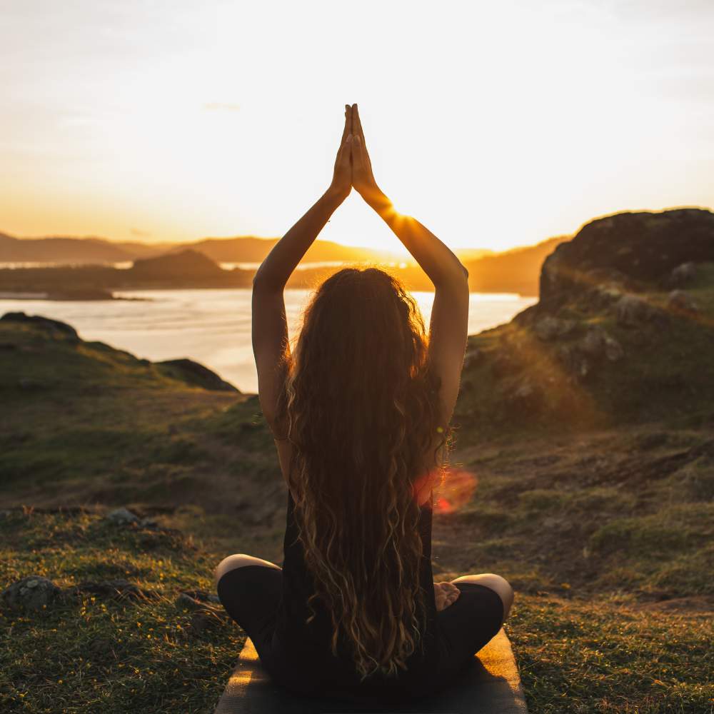 Femme assise en position de méditation face au coucher du soleil, mains jointes au-dessus de la tête, symbolisant la sérénité, le bien-être et l’équilibre intérieur apportés par les gummies au CBD.