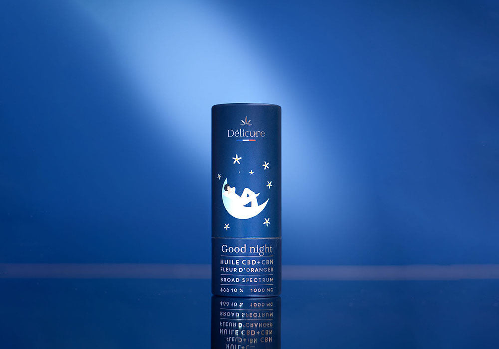 Image d'un flacon de produit Délicure intitulé Good night, conçu pour favoriser le sommeil. Le flacon est cylindrique et de couleur bleue, avec une illustration représentant une personne dormant sur un croissant de lune, entourée d'étoiles. Le texte sur l'étiquette indique que le produit contient de l'huile CBD et CBN avec de la fleur d'oranger, et affiche une concentration de 1000 mg à large spectre.
