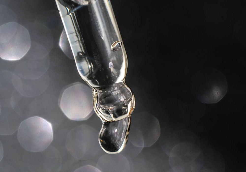Image d'un compte-gouttes en verre avec une goutte d'huile de cbd sport et repair, qui réduit les douleurs liées aus muscles et articulations, transparent suspendue à son extrémité. L'arrière-plan est sombre avec des reflets lumineux flous qui créent un effet de bokeh, mettant en valeur la clarté et la pureté du liquide. La composition de l'image évoque des thèmes de précision et de soin, souvent associés à des produits de bien-être ou de beauté.