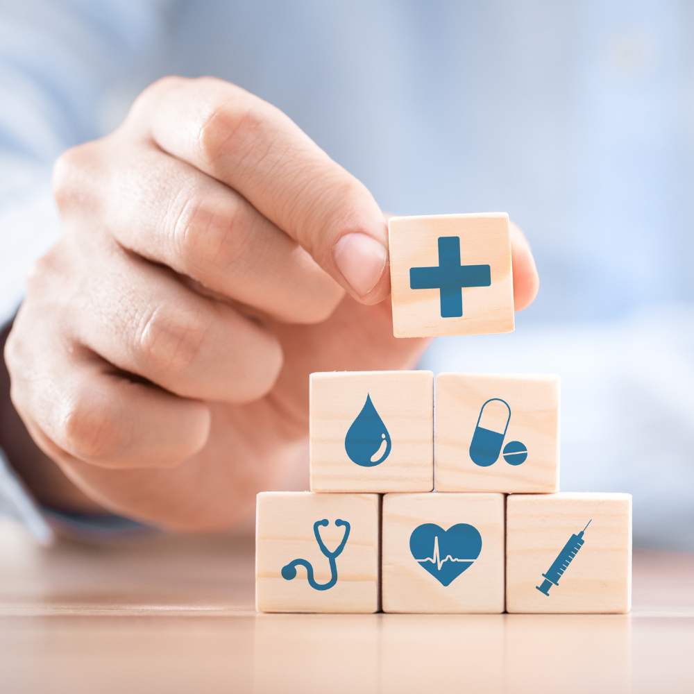 Main tenant un cube en bois avec une croix médicale bleue, posant ce cube au sommet d’une pyramide de blocs portant des symboles de santé (stéthoscope, cœur, pilule, goutte, seringue), représentant la sécurité, la légalité et le contrôle médical autour du CBD.