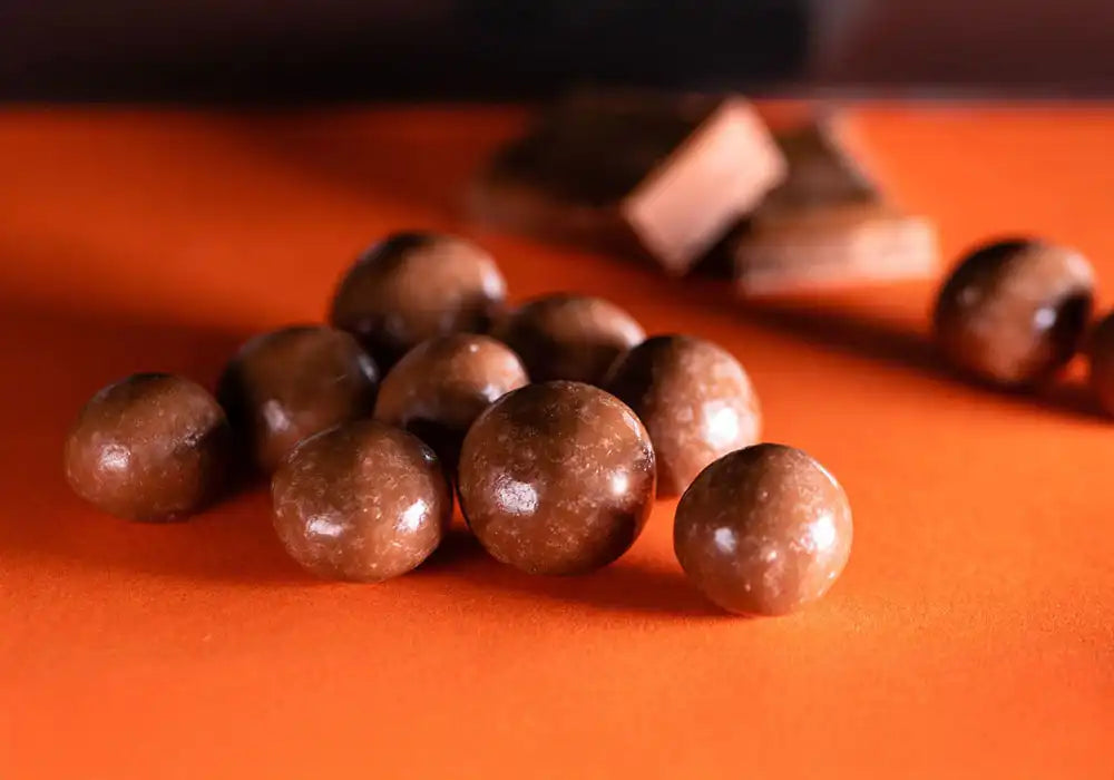 Image de gros plan sur des bonbons au chocolat au CBD de la marque Délicure. Ces bonbons ronds et brillants sont disposés sur une surface orange, avec des morceaux de chocolat en arrière-plan. Chaque bonbon est infusé avec du CBD, conçu pour aider à soulager le stress et promouvoir la relaxation. La présentation met en valeur la richesse et la texture lisse du chocolat, soulignant la combinaison délicieuse et apaisante de ce produit contre le stress.