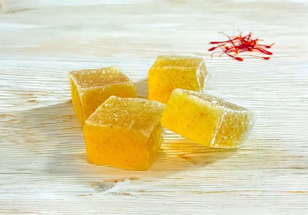 Image de quatre gummies jaunes avec des pigments rouges posés sur une surface en bois clair. À côté des gummies, il y a côté des gummies, des filaments de safran rouge. La texture des gummies semble douce et qualitative, suggérant une consistance moelleuse. La disposition et les couleurs vives des ingrédients créent une présentation appétissante et naturelle.