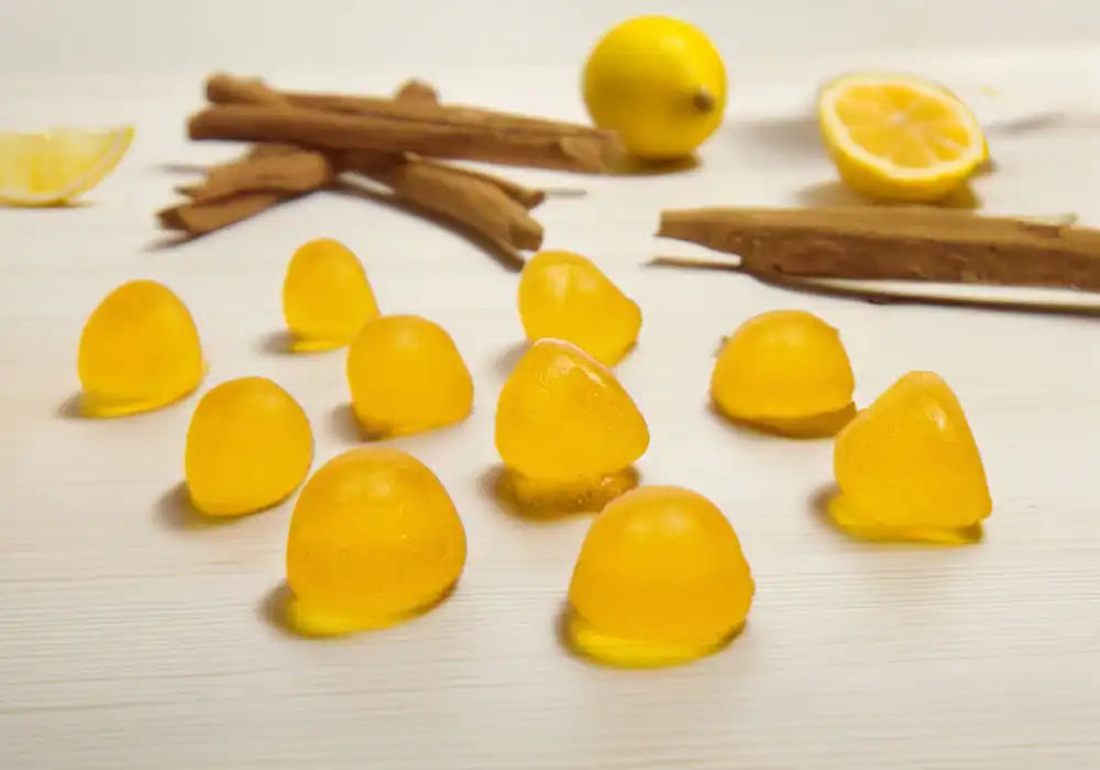 Image de gros plan sur des gummies au CBD de la marque Délicure, saveur citron. Ces gummies jaunes, présentés sur une surface claire, sont entourés de morceaux de citron frais et de bâtons de cannelle, mettant en valeur leur composition naturelle. Chaque gummy contient 10 mg de CBD, conçu pour aider à soulager le stress. La photo met en avant la texture douce et l'arôme rafraîchissant du citron, soulignant l'efficacité apaisante de ces compléments alimentaires.