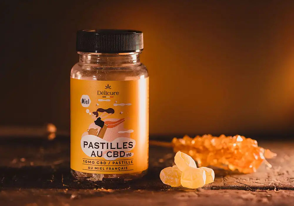 Image d'un flacon de pastilles au CBD de la marque Délicure, saveur miel. Le flacon est transparent avec un couvercle noir, et l'étiquette est jaune. L'illustration sur l'étiquette montre une personne savourant une boisson chaude. Le texte indique que chaque pastille contient 10 mg de CBD et que le produit est fabriqué avec du miel français, avec un total de 40 pastilles par flacon. À côté du flacon, on peut voir quelques pastilles au CBD et un bâtonnet de miel cristallisé.