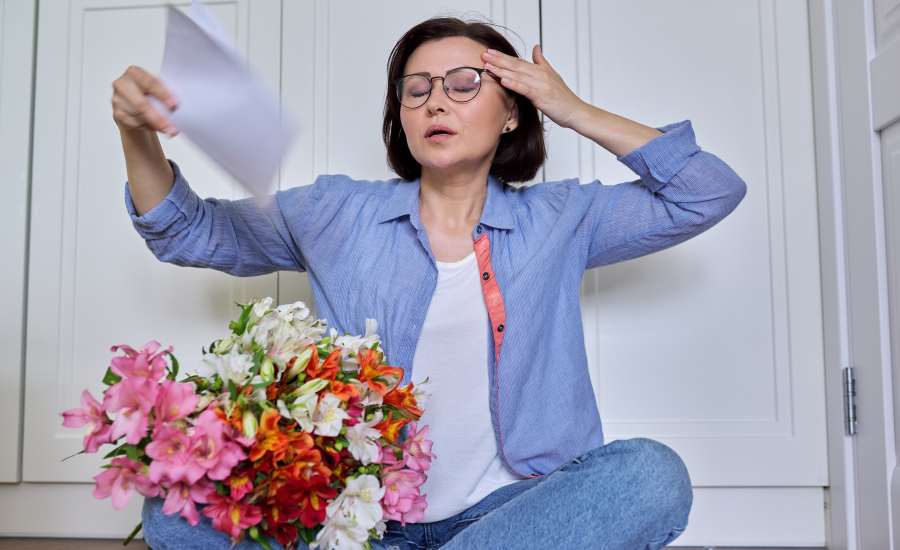Femme d’âge moyen assise au sol, ventilant son visage avec une feuille de papier pour illustrer une bouffée de chaleur de la ménopause