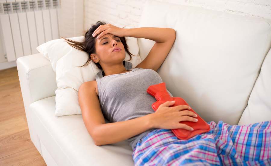 Femme allongée sur un canapé, une bouillotte chaude posée sur le bas-ventre pour apaiser les règles douloureuses