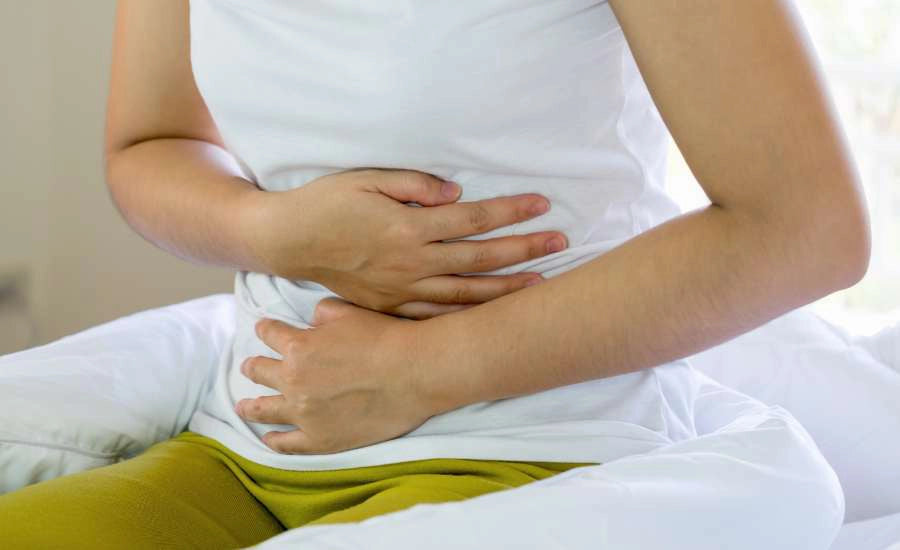 Donna seduta su un letto, con indosso una maglietta bianca e pantaloni verdi, con le mani appoggiate sull'addome in segno di dolore, sintomo di problemi digestivi (gonfiore, bruciore di stomaco, stitichezza).
