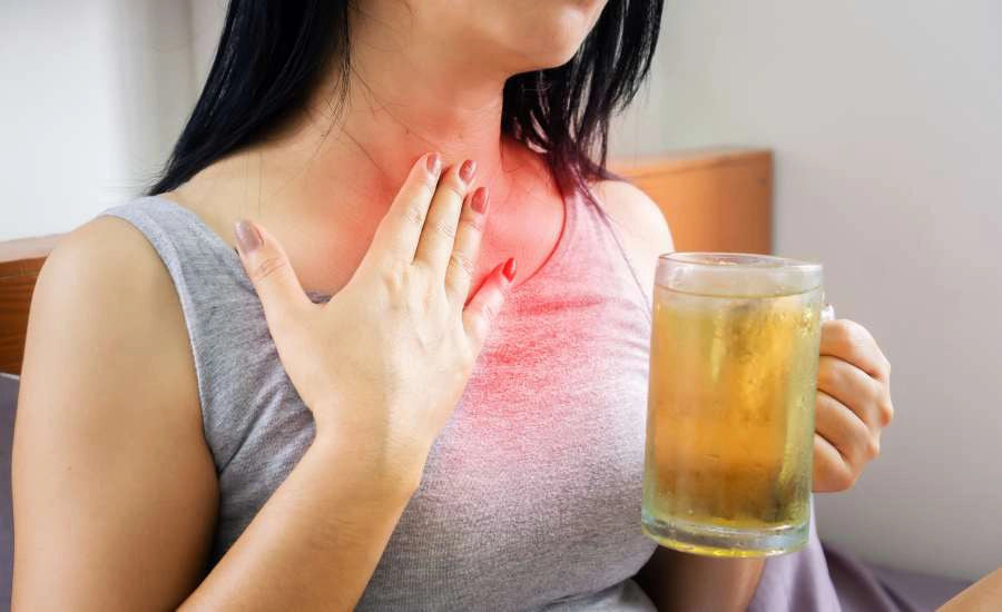 Primo piano di una donna seduta con una canottiera grigia, che tiene un boccale di birra nella mano destra e si mette la mano sinistra sulla gola e sul petto; Un velo rosso all’altezza dello sterno simboleggia il bruciore di stomaco.